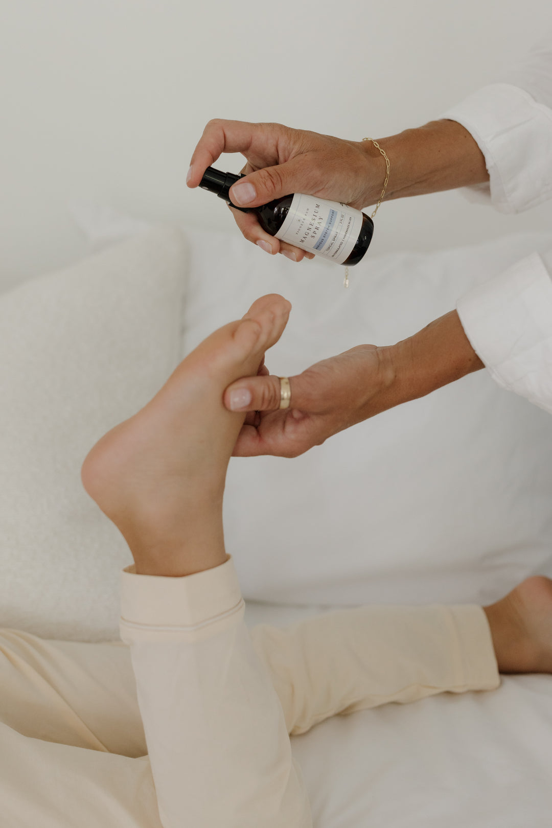 Hands spraying Pioneer Pam Magnesium Oil Spray onto a bare foot near a bed