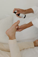 Hands spraying Pioneer Pam Magnesium Oil Spray onto a bare foot near a bed