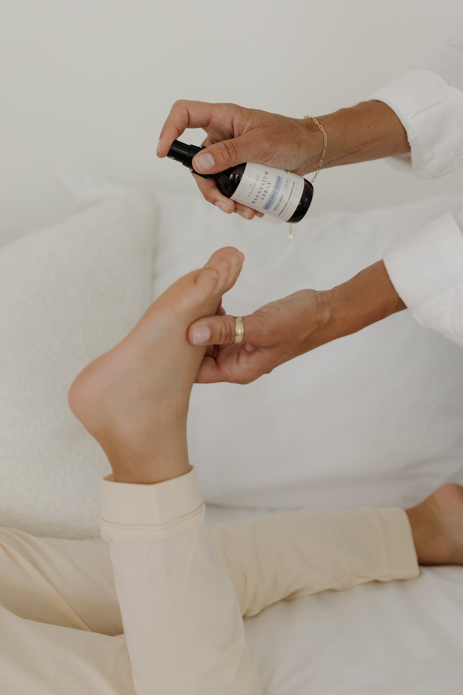 Hands spraying Pioneer Pam Magnesium Oil Spray onto a bare foot near a bed