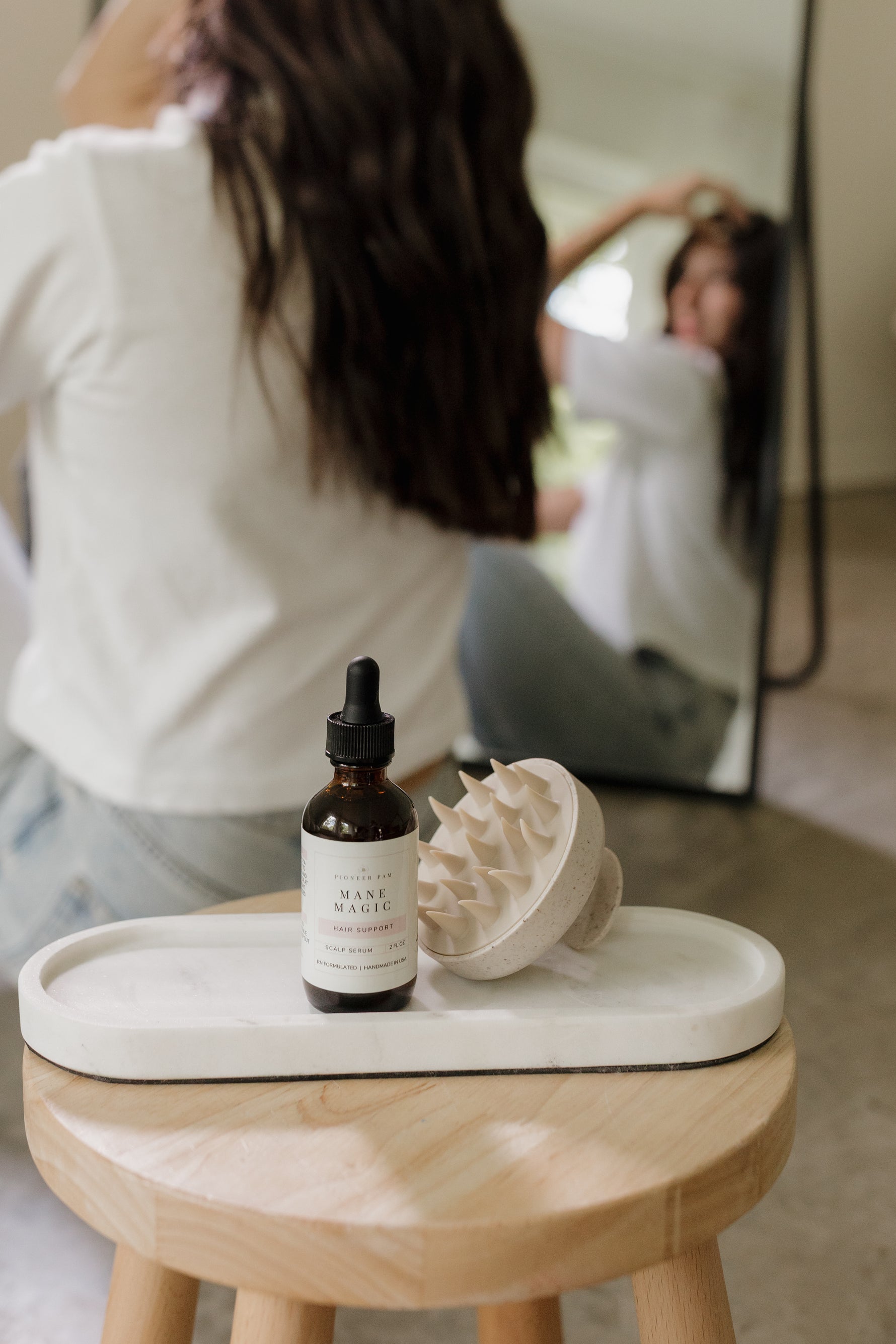 Woman styling her hair at a mirror with Pioneer Pam Mane Magic bottle and hairbrush nearby