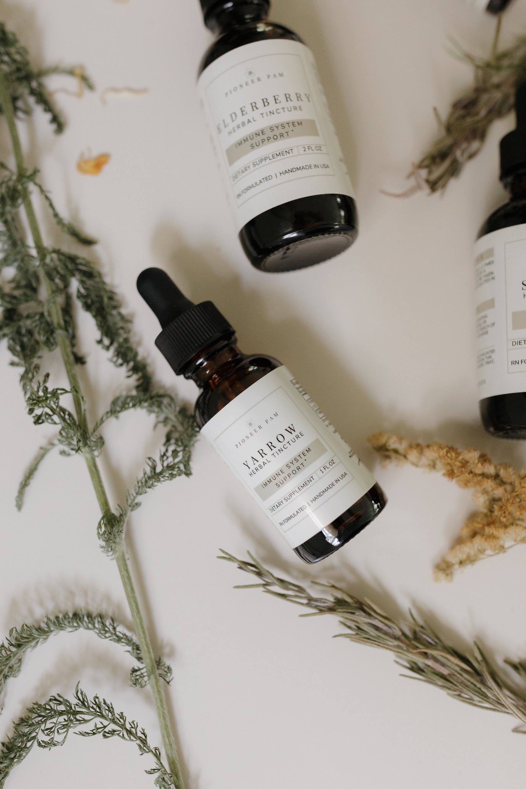 Flat lay of Pioneer Pam herbal tincture bottles, including Yarrow, surrounded by fresh and dried botanical sprigs on a light background.