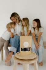 Mother embracing her children next to the Pioneer Pam DIY Elderberry Syrup Kit, set against a sage green background.