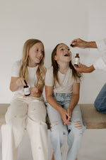 Two children being given herbal tincture drops by an adult, using small amber dropper bottles